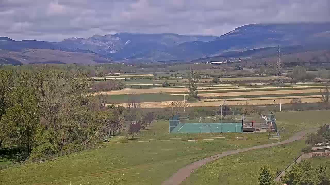 A green sports court sits in a grassy field, overlooking a valley of cultivated land and distant mountains under a partly cloudy sky.