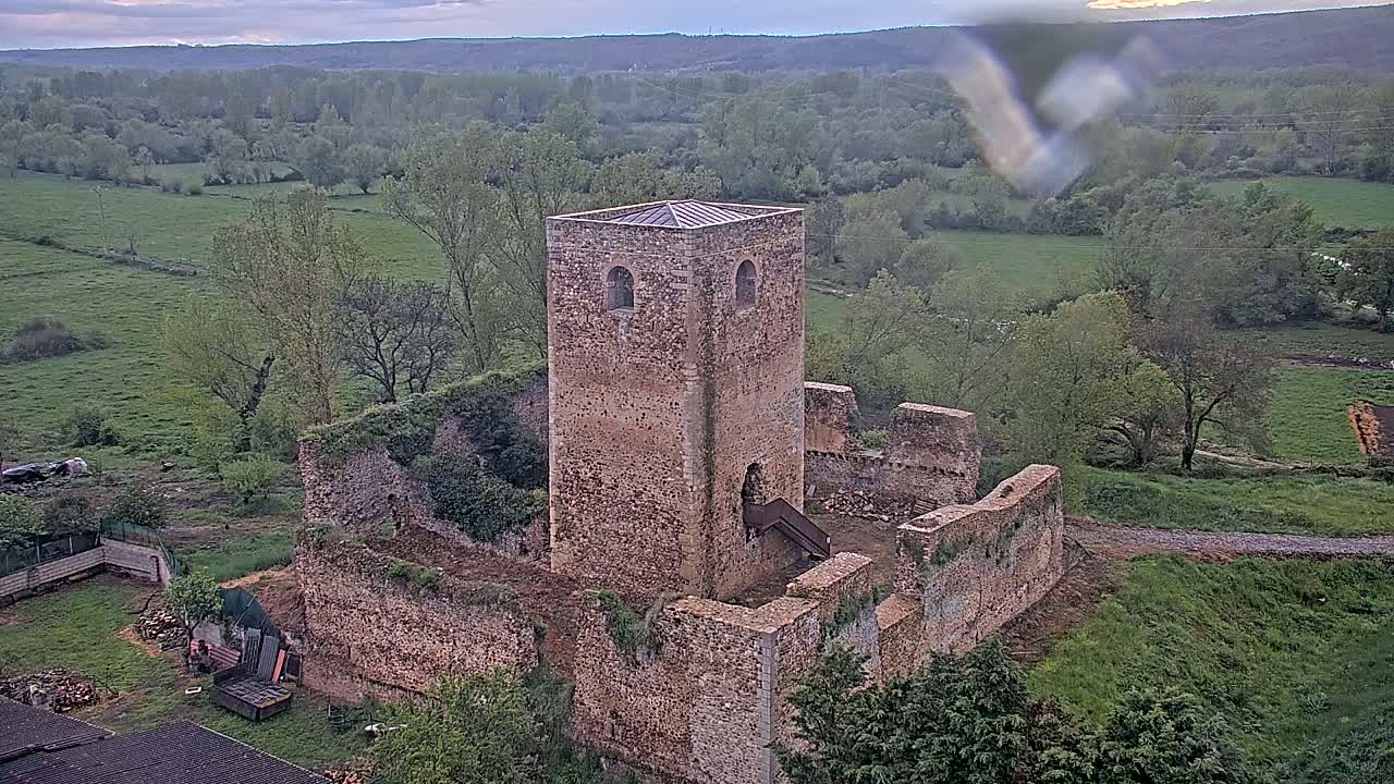 A stone tower and ruined walls of a castle sit atop a verdant hill overlooking a valley of green fields and trees under a cloudy sky.