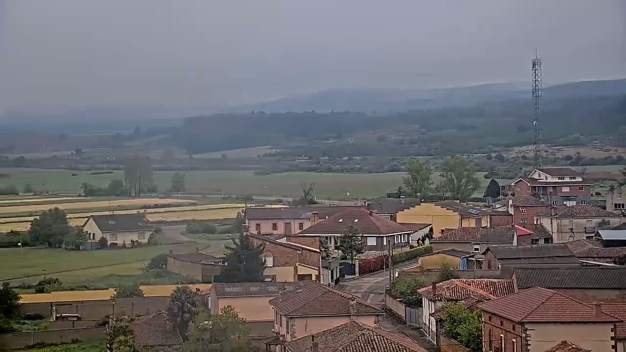 A hazy, overcast day reveals a sprawling village nestled in a valley, surrounded by fields and distant hills.