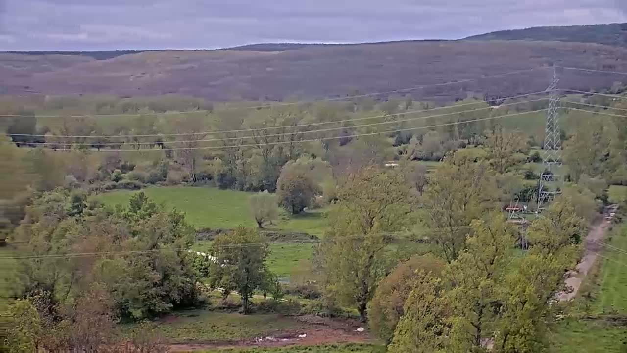 A high-angle view shows a pastoral valley under a cloudy sky, with green fields, scattered trees, and power lines crisscrossing the landscape.