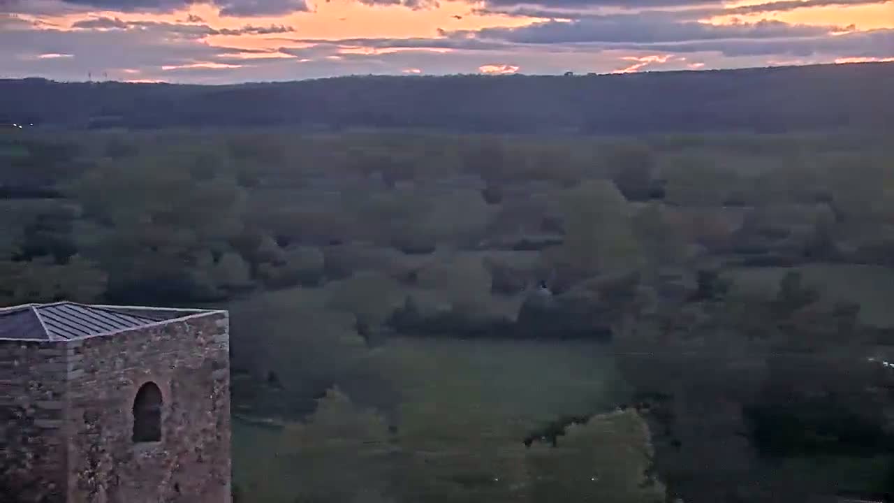 A stone tower with a metal roof is situated atop a hill overlooking a vast expanse of wooded land under a dusky, sunset sky.