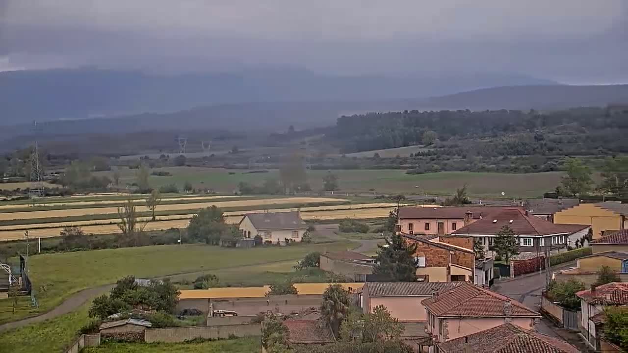 A village with red-tiled roofs sits in a valley under an overcast sky, with fields and distant mountains visible in the background.