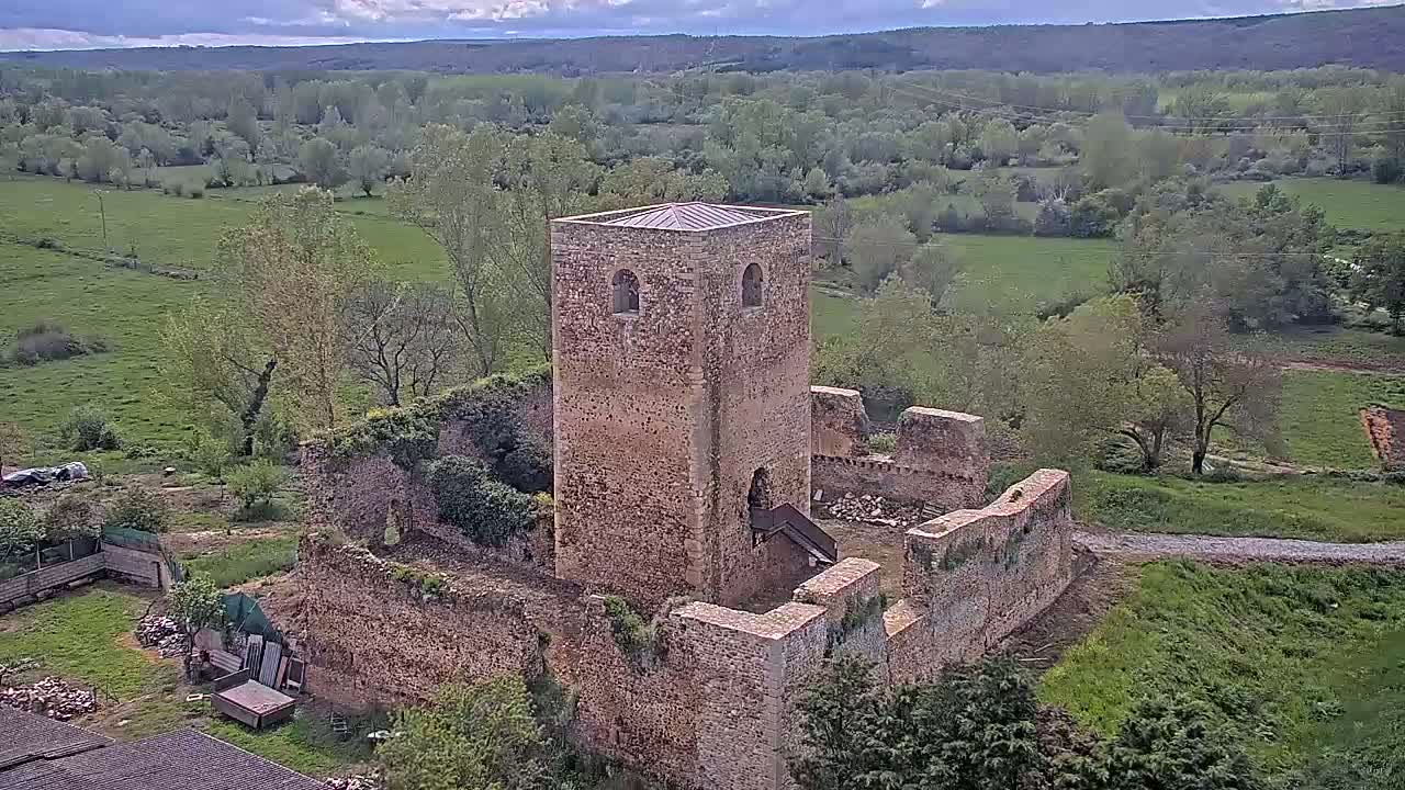 An aerial view shows the stone ruins of a tower and castle walls surrounded by green fields and trees under a partly cloudy sky.