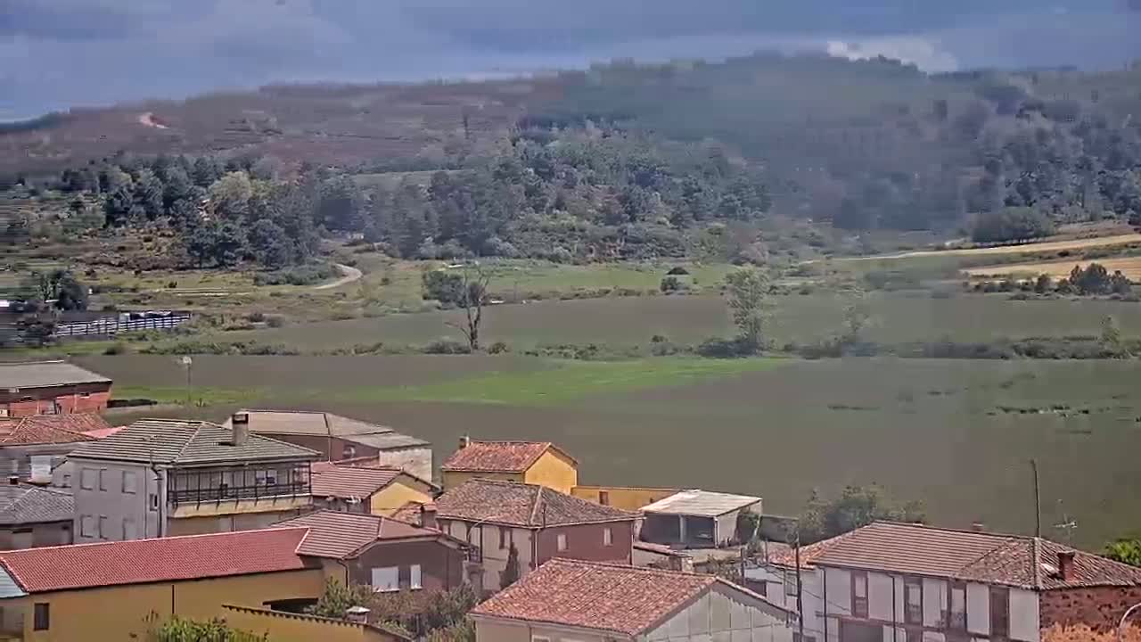 A village of red-tiled houses sits nestled in a valley under a partly cloudy sky, overlooking a flat expanse of land and a line of trees in the distance.