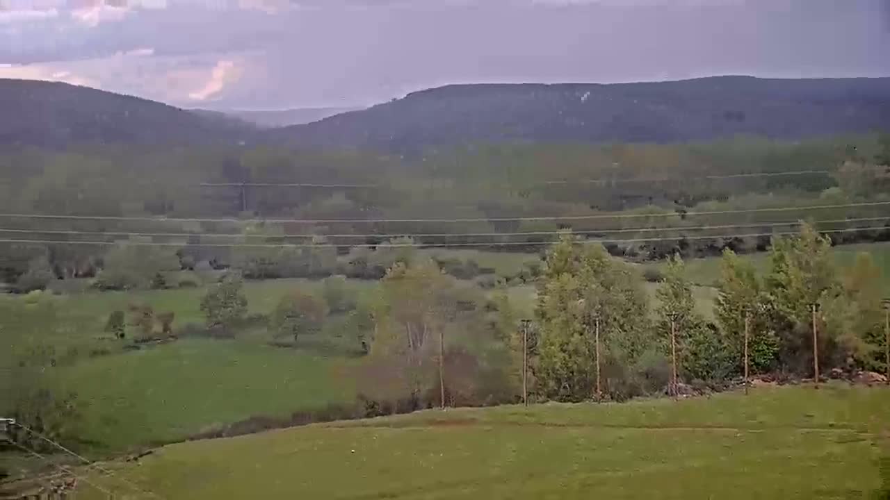 A sprawling green valley dotted with trees and fields stretches toward distant, hazy mountains under a partly cloudy sky.