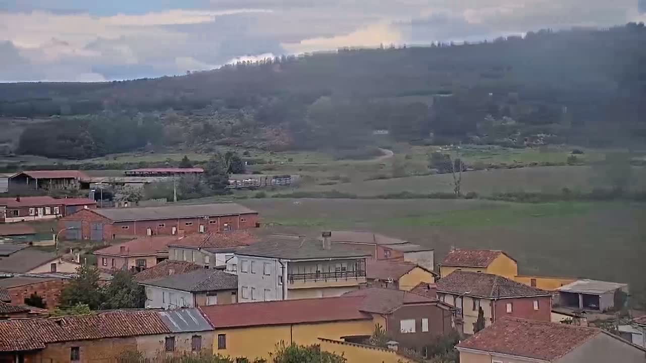 A small village of red-tiled houses sits in a valley under a cloudy sky, with a forested hill visible in the background.