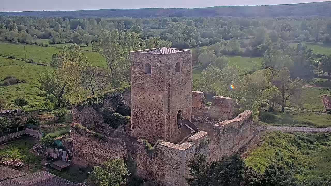 An aerial view shows the stone ruins of a castle, including a tall, intact tower, set in a verdant landscape on a sunny day.