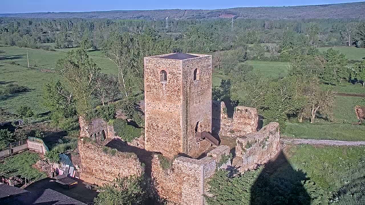 An aerial view shows the stone ruins of a castle, including a tall, intact tower, situated in a verdant landscape under a clear sky.