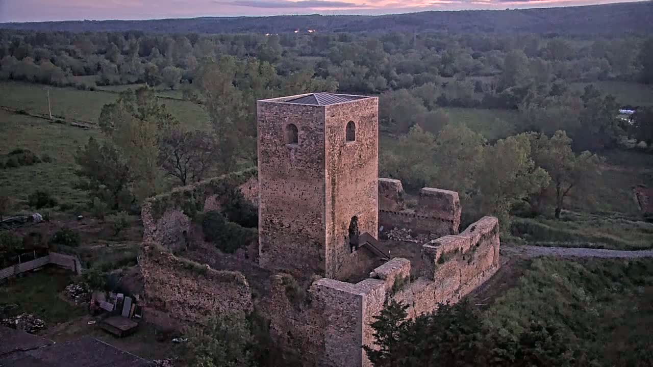 Stone castle ruins, including a tall standing tower, are situated on a hill overlooking a verdant valley under a twilight sky.