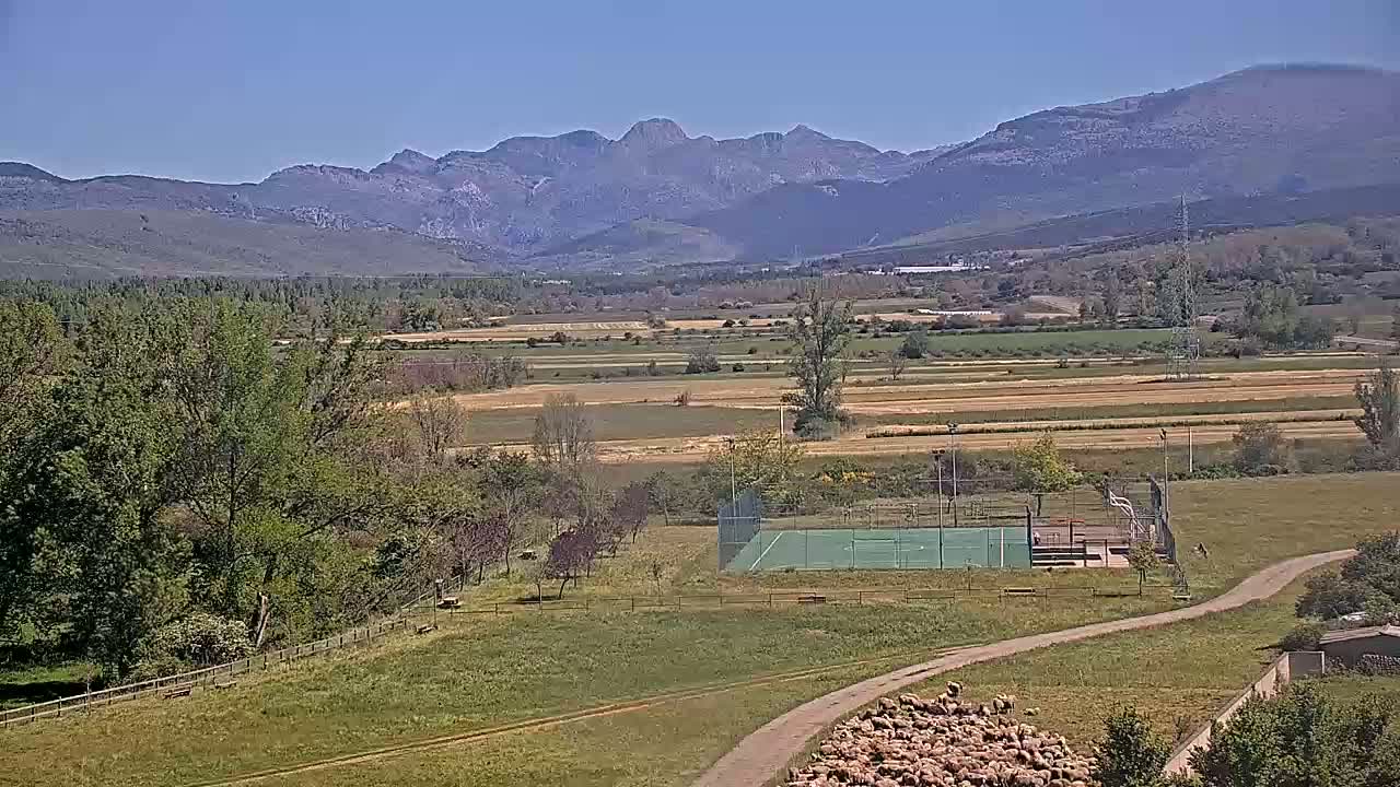 A green valley with a fenced-in sports court, a flock of sheep, and a mountain range in the background under a clear blue sky.