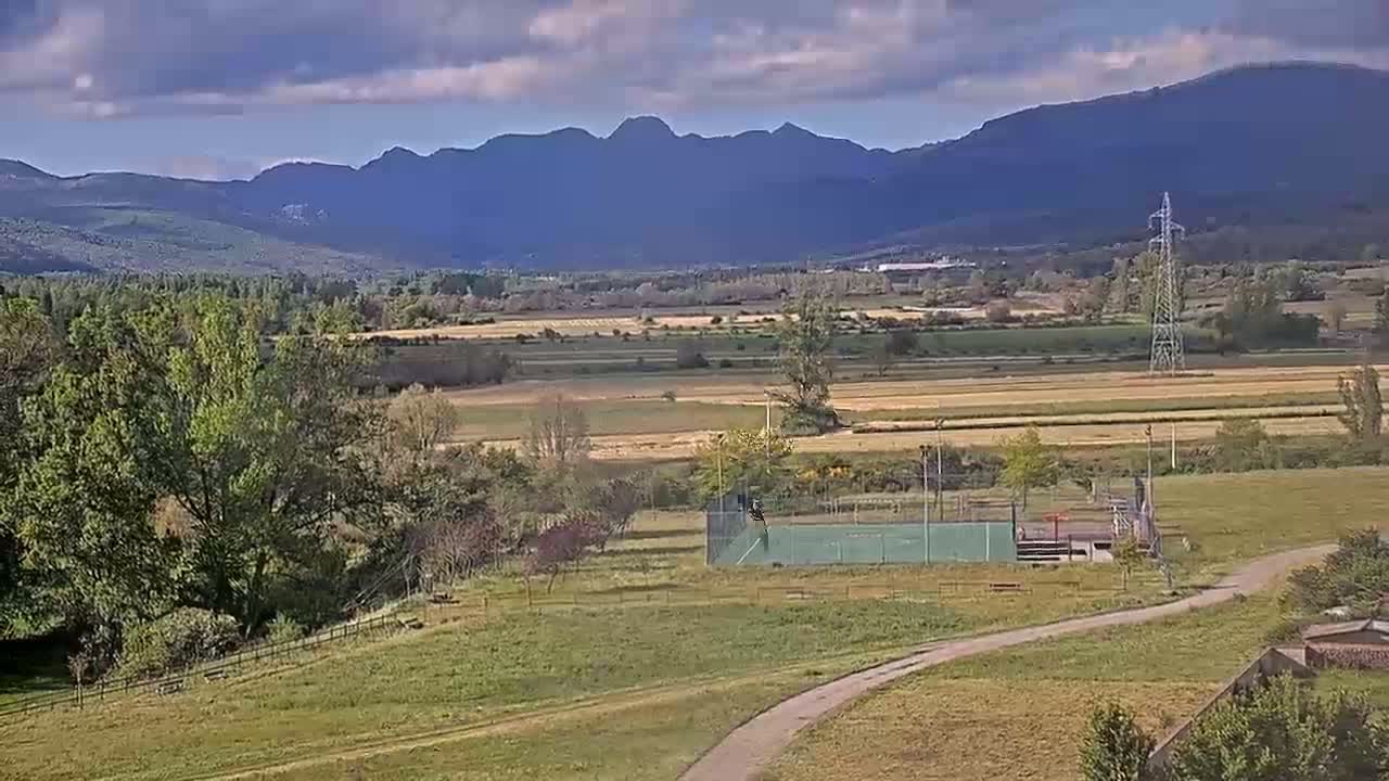 A partly cloudy day reveals a valley with fields, trees, a tennis court, and distant mountains.