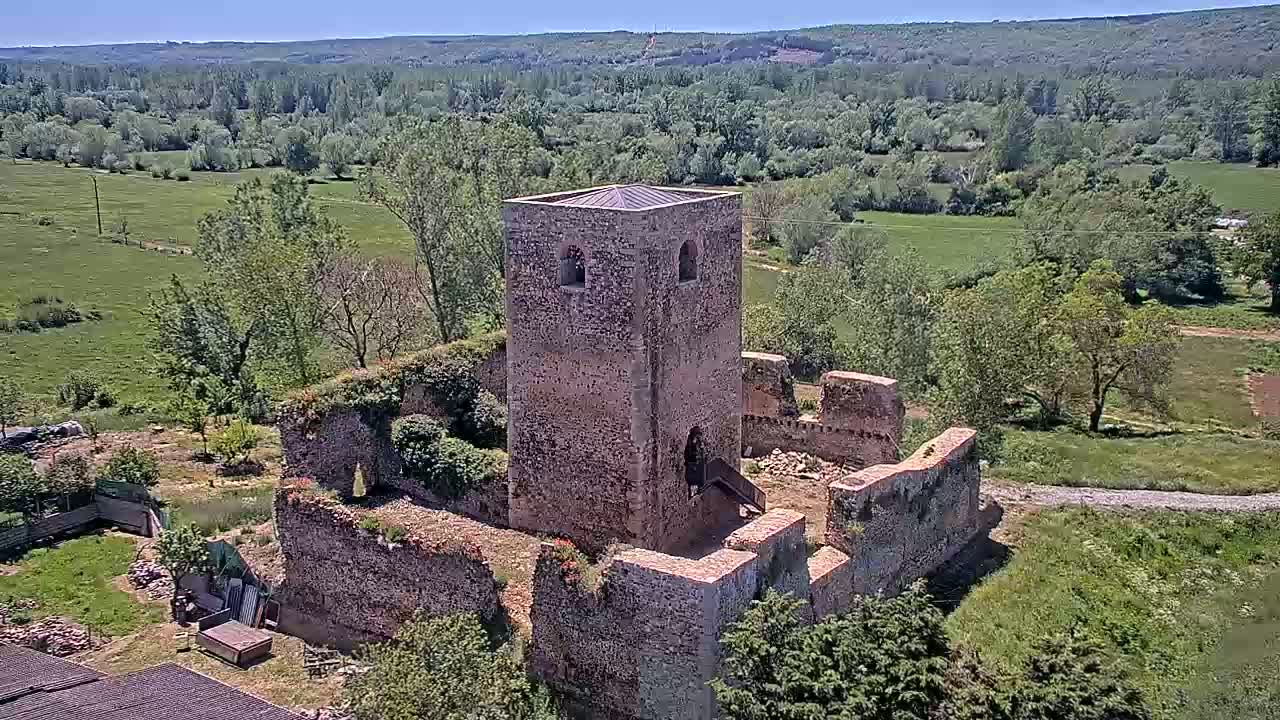 An aerial view shows the ruins of a stone tower and surrounding walls on a sunny day, overlooking a verdant valley.
