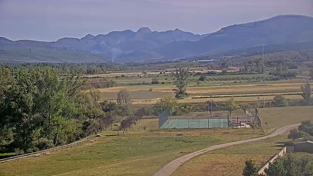 A green sports court sits in a rural landscape under a mostly clear sky, with distant mountains visible in the background.