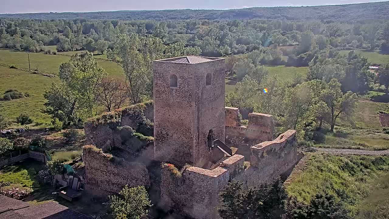 An aerial view shows the stone ruins of a castle on a sunny day, surrounded by green fields and trees.