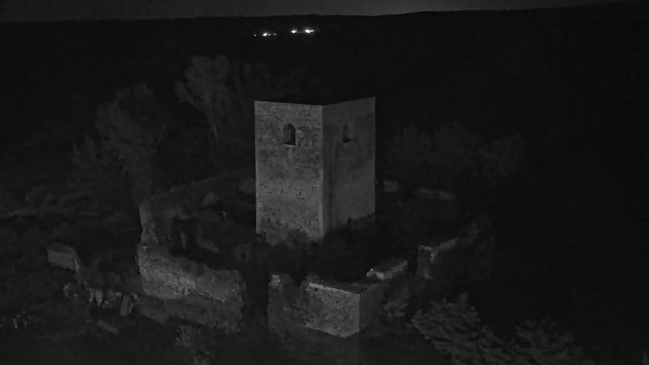 An aerial nighttime view shows a stone tower and surrounding ruins, with a dark, possibly wooded landscape in the background under a clear, dark sky.