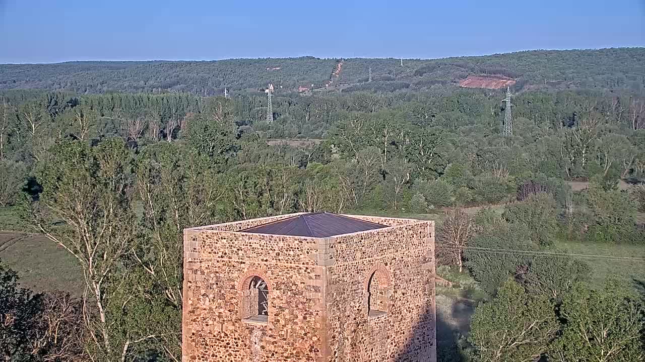 A stone tower with arched windows sits atop a hill overlooking a vast expanse of green forest under a clear blue sky.