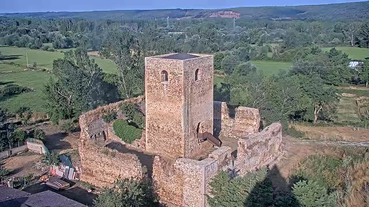 An aerial view shows the ruins of a stone tower and surrounding walls, situated in a verdant landscape under a clear, sunny sky.