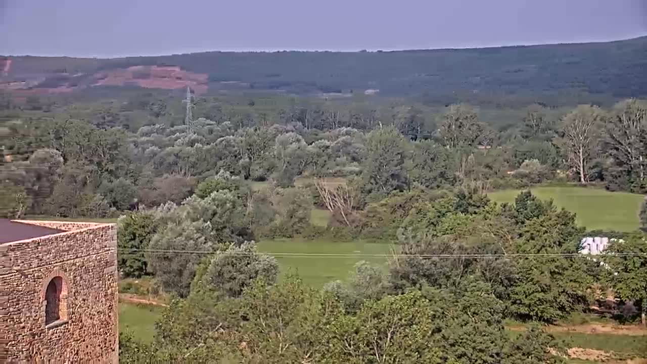 A stone structure with an arched opening overlooks a verdant valley dotted with trees under a clear sky.