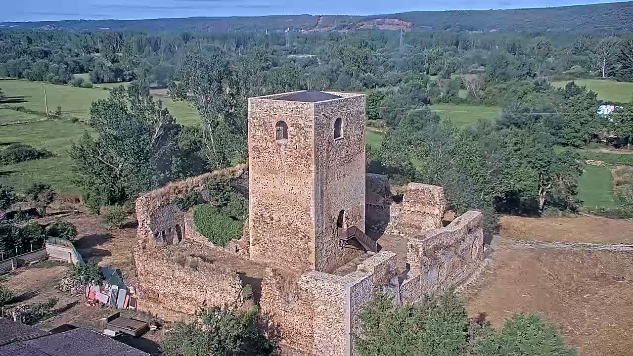 An aerial view shows the ruins of a stone tower and surrounding walls on a sunny day, overlooking a verdant valley.