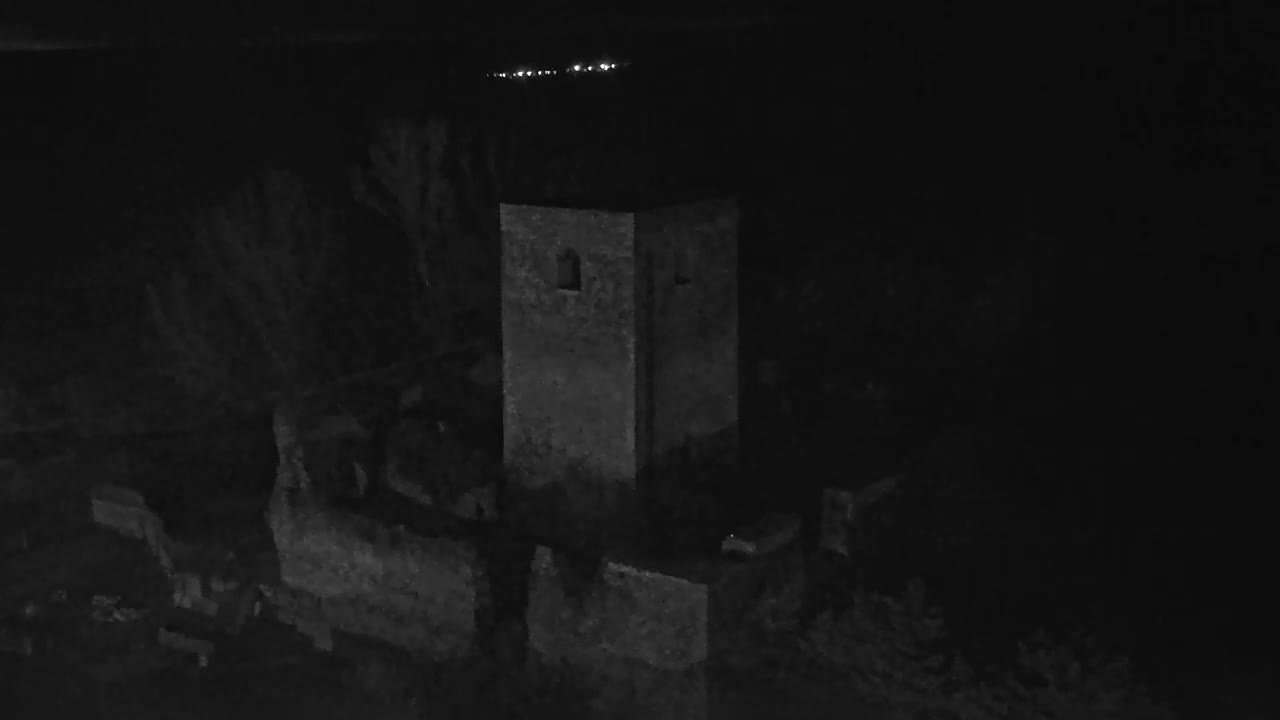 A stone tower and its surrounding ruins are dimly visible at night, with a distant line of lights on the horizon under a clear sky.