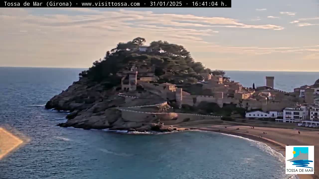 A coastal town with a medieval-style castle perched atop a rocky hill overlooking a calm, blue sea under a partly cloudy sky.