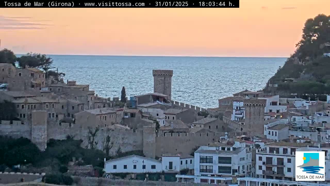 A coastal town with stone buildings and a tower overlooks a calm sea under a sunset sky.