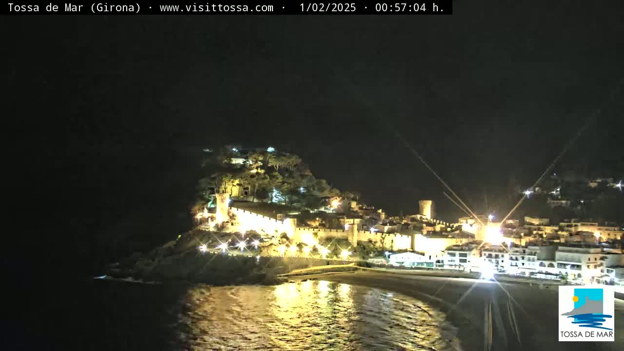 A nighttime coastal town with illuminated buildings and walls sits on a hill overlooking a dark beach and calm water.
