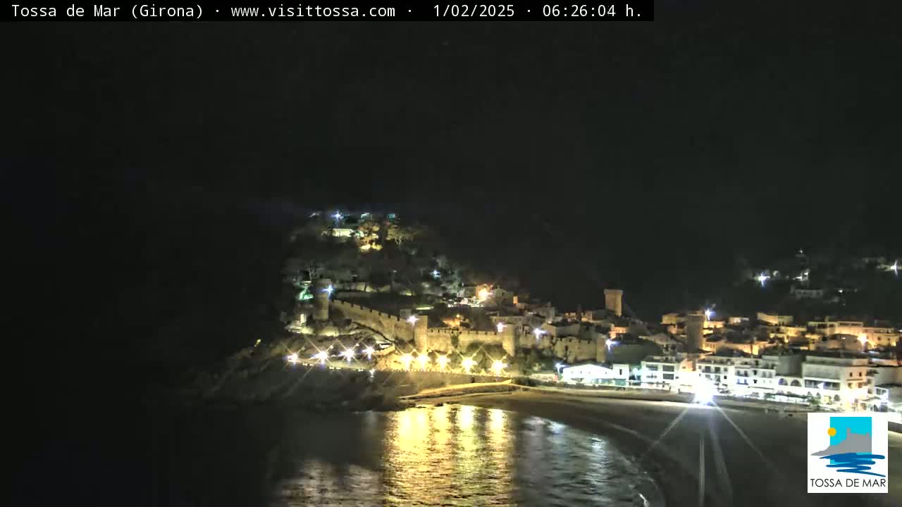 A nighttime coastal town with a walled fortress illuminated on a hill overlooking a calm, dark bay.