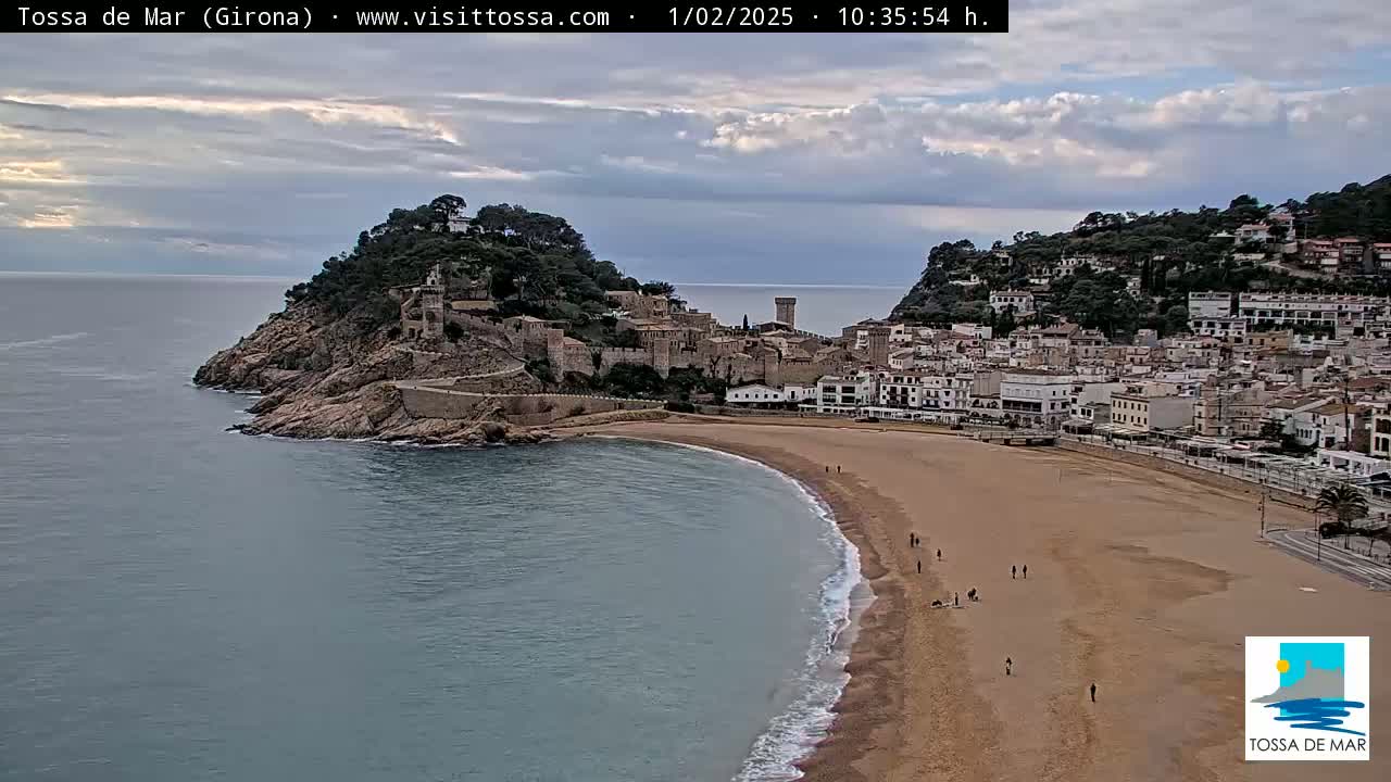 A coastal town with a sandy beach and a medieval castle sits on a hill overlooking a calm sea under a partly cloudy sky.
