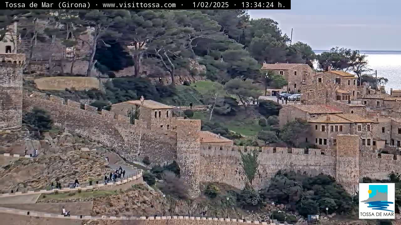 An overcast day reveals a coastal town with stone buildings and walls, nestled on a hillside overlooking the sea.
