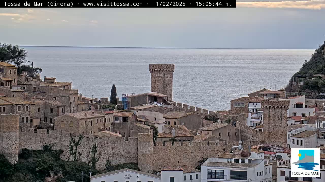 A stone walled coastal town with a prominent tower overlooks a calm sea under a partly cloudy sky.