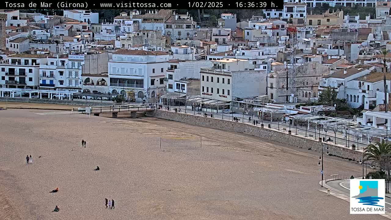 A mostly empty beach stretches before a coastal town of white buildings under a partly cloudy sky.