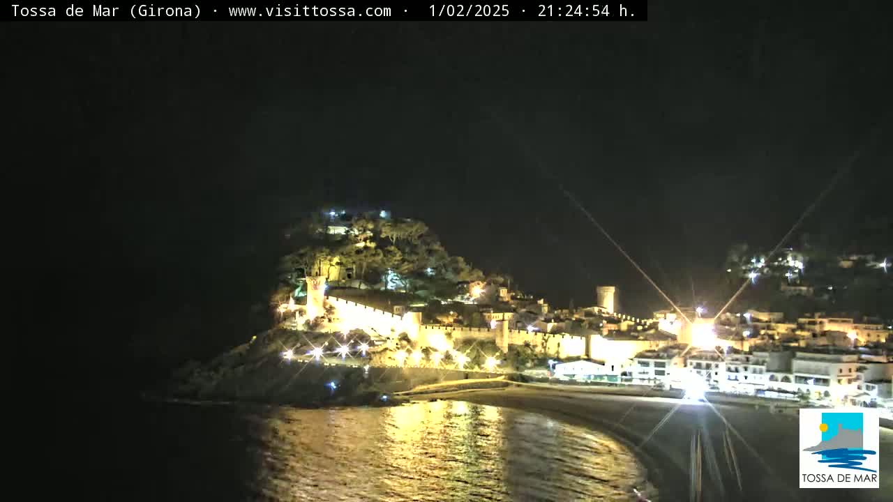 A nighttime coastal town, illuminated with lights, is situated on a hill overlooking a calm, reflective body of water.