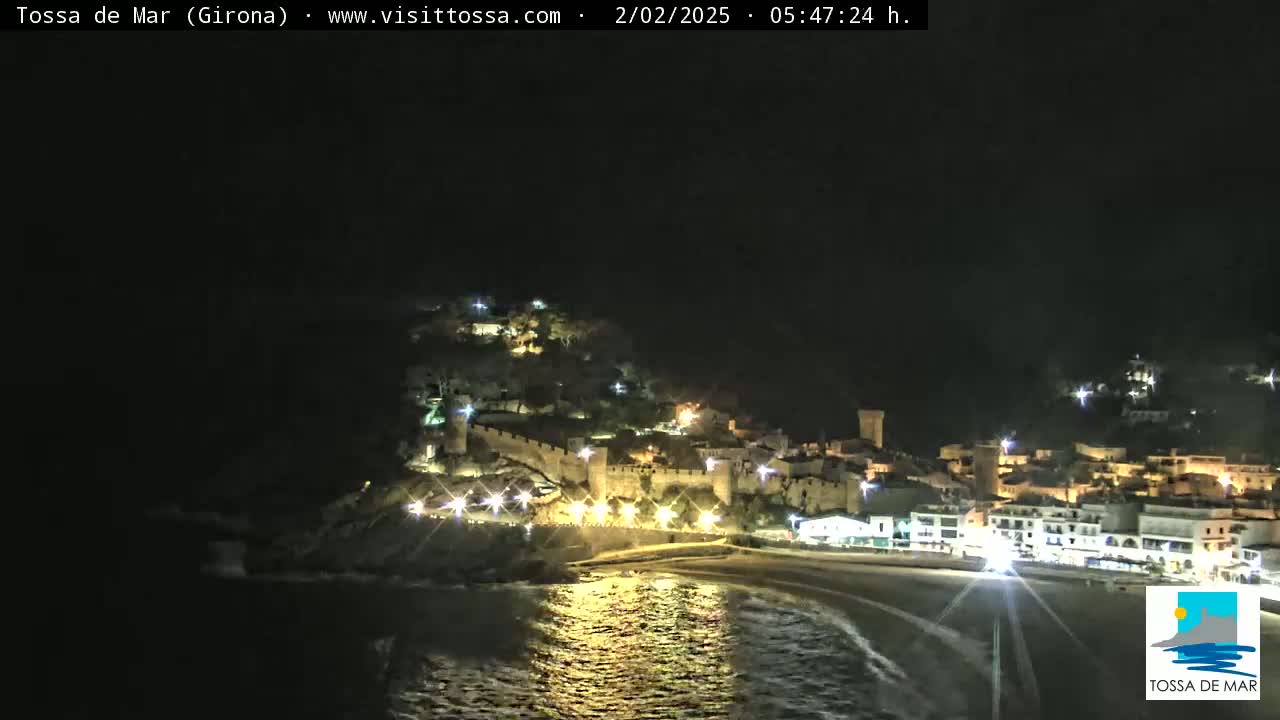 A nighttime coastal town with a walled fortress is illuminated, its lights reflecting on the dark water.