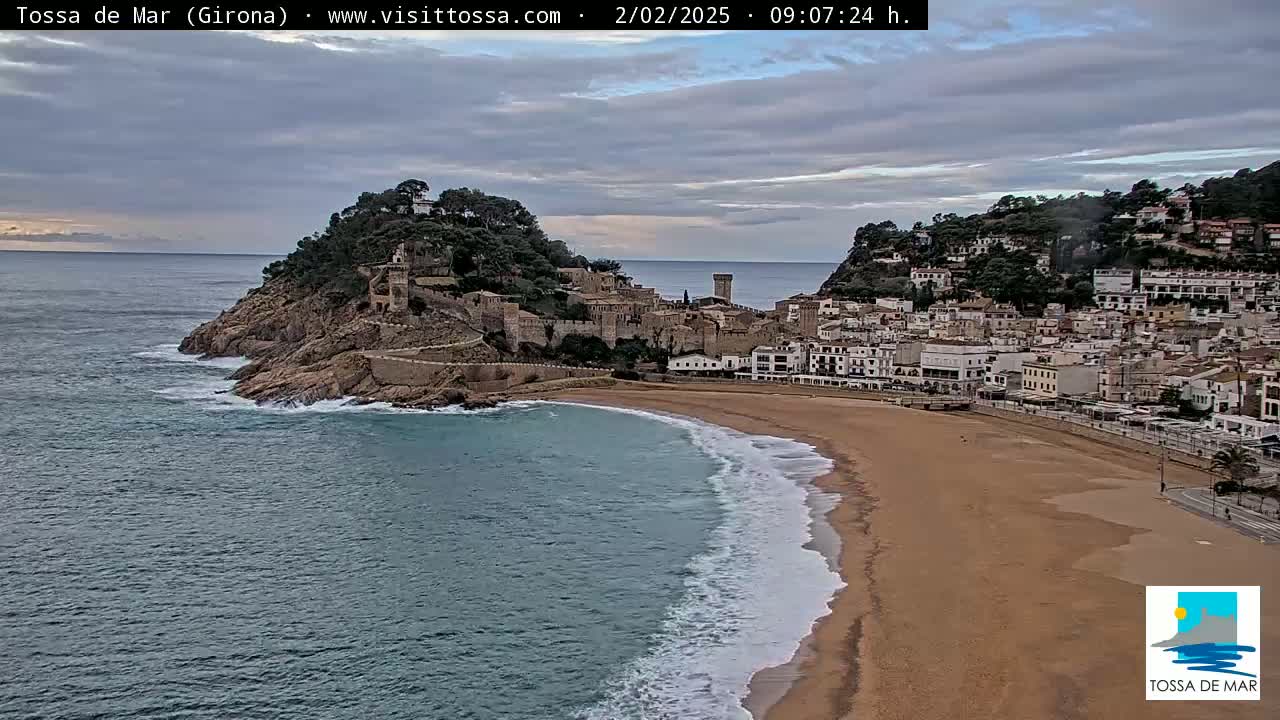 A coastal town with a medieval castle sits on a hill overlooking a sandy beach with gentle waves under a cloudy sky.