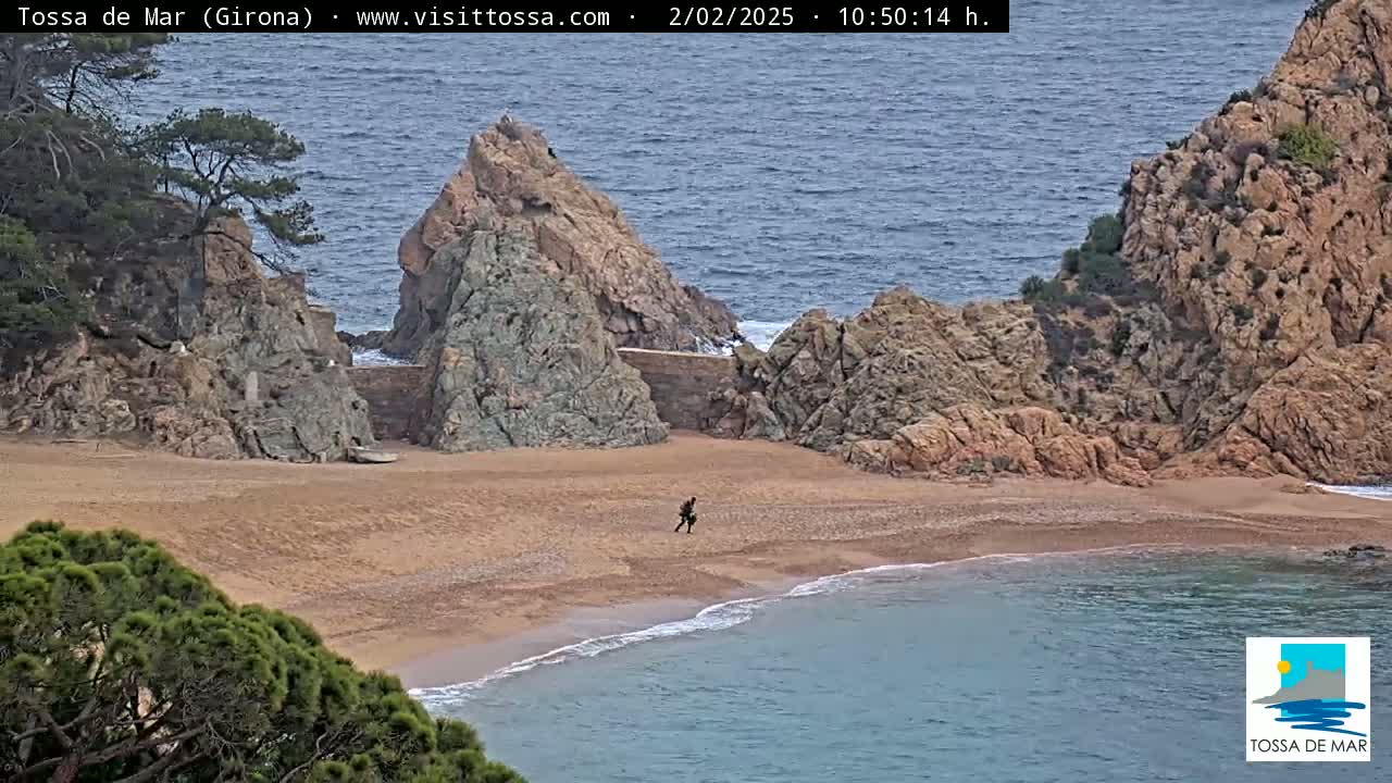 A sandy cove nestled between rocky cliffs, with a person walking on the beach, under an overcast sky.