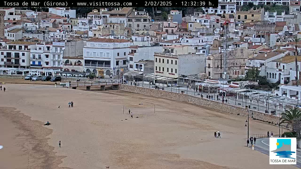 A mostly empty sandy beach stretches below a coastal town of white buildings under an overcast sky.