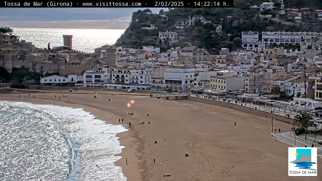 A sandy beach with gentle waves laps the shore in front of a coastal town built on a hillside under a partly cloudy sky.
