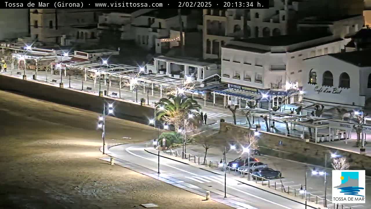 A nighttime view of a seaside town's illuminated promenade and parking area with several cars and a few pedestrians.