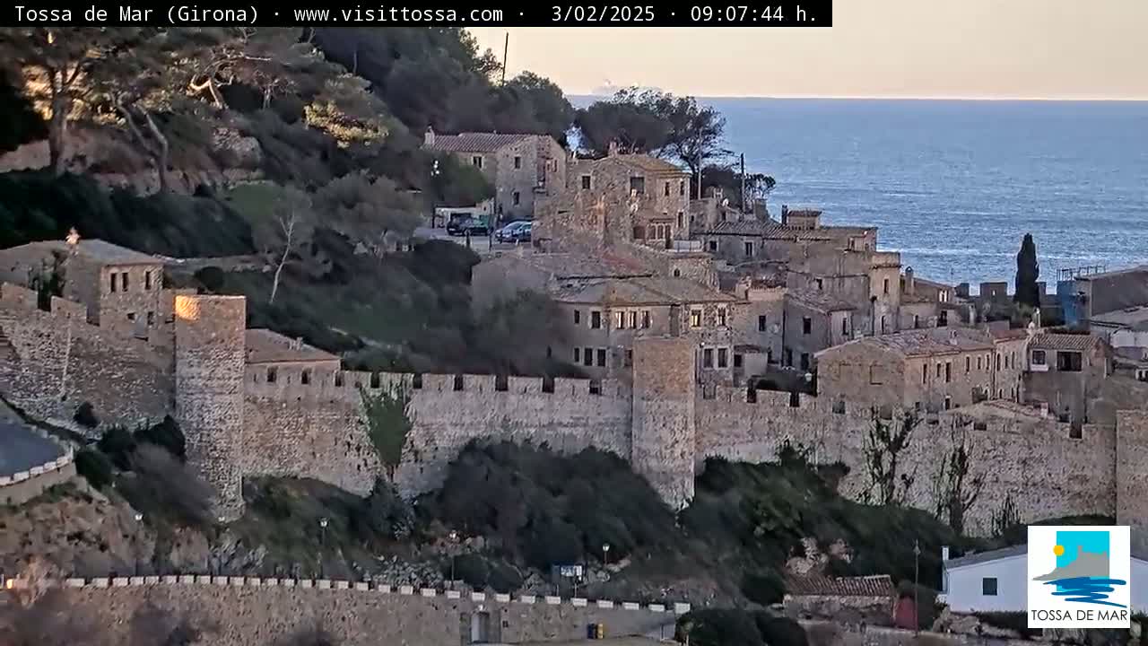 A stone walled medieval town overlooks a calm sea under a clear, twilight sky.