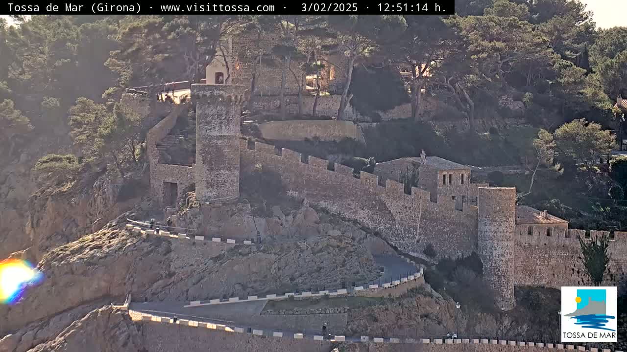 A stone wall encircles a hilltop town, winding roads lead up to the fortifications, and the weather appears sunny.
