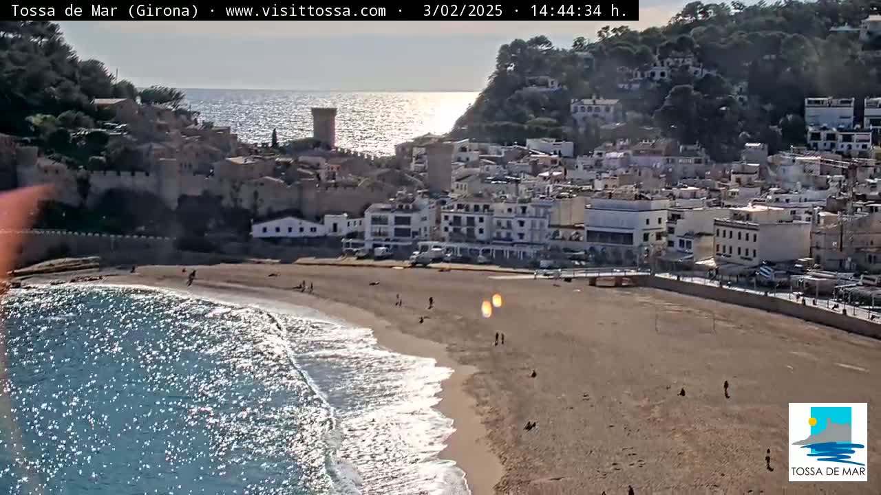 A sandy beach with a few people on it borders a town of white buildings nestled against a hillside overlooking a sparkling blue sea under a bright, sunny sky.