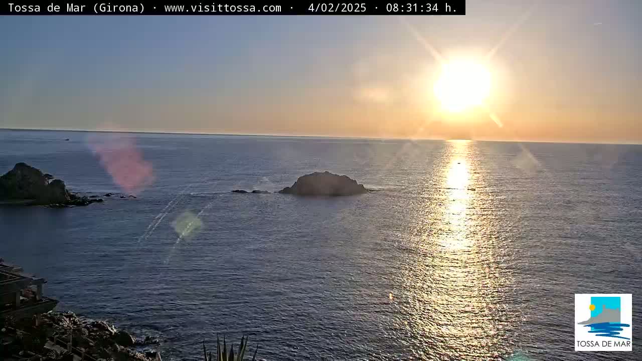 A bright sun shines over a calm ocean with two small rocky islands visible in the foreground during what appears to be sunrise or sunset.