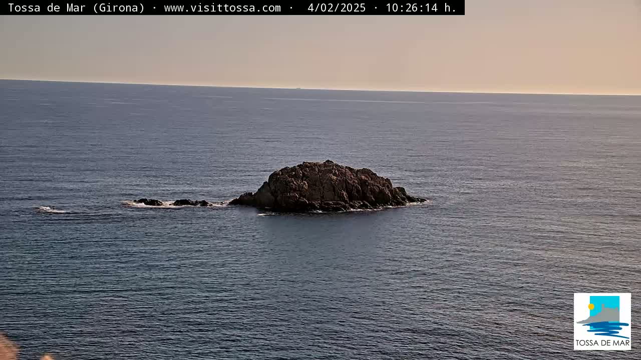 A small, rocky island sits in calm, dark-blue ocean water under a clear, pale sky.