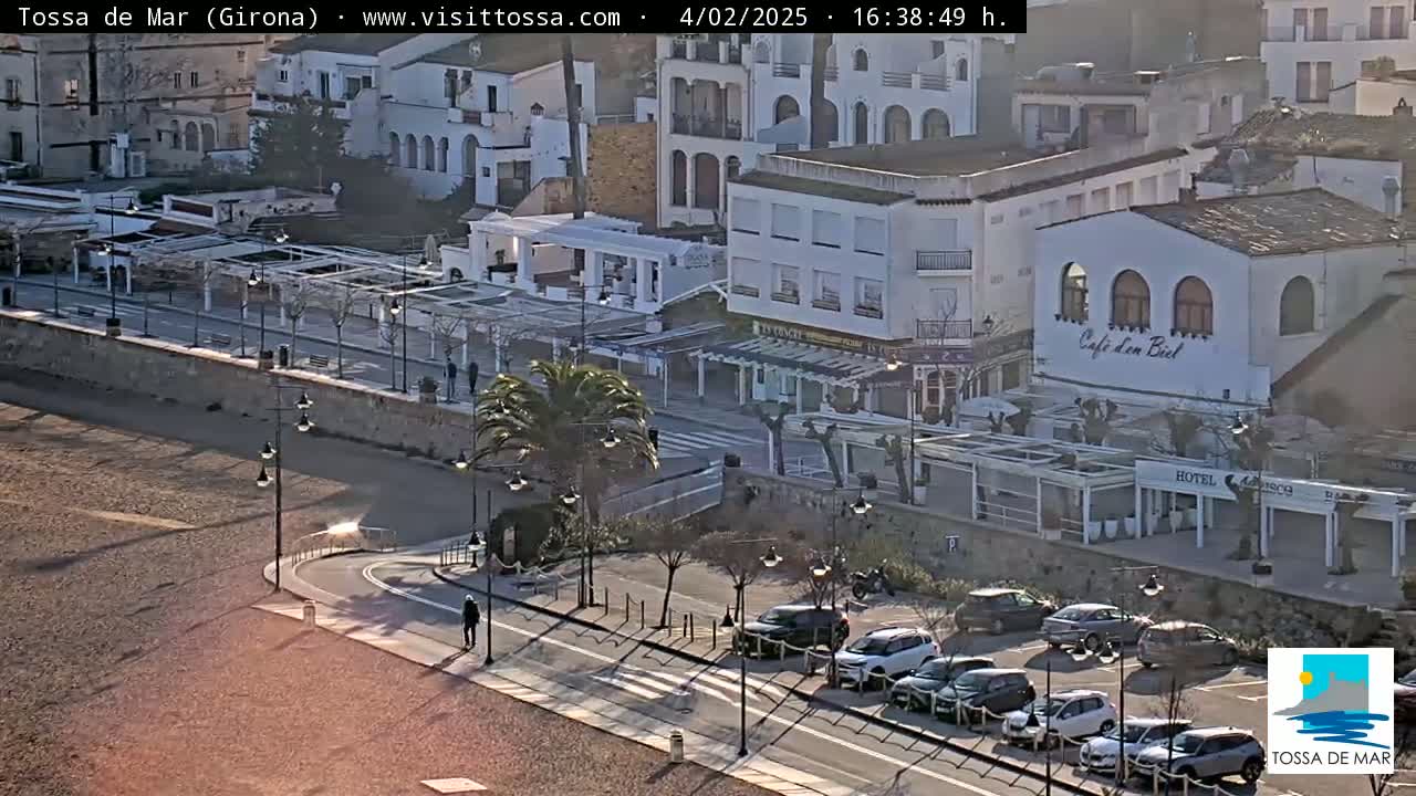 A sunny coastal town with white buildings, a sandy beach, and parked cars along a road.