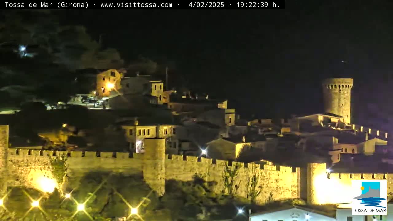 A nighttime view of a walled, illuminated town with a prominent tower, under clear skies.