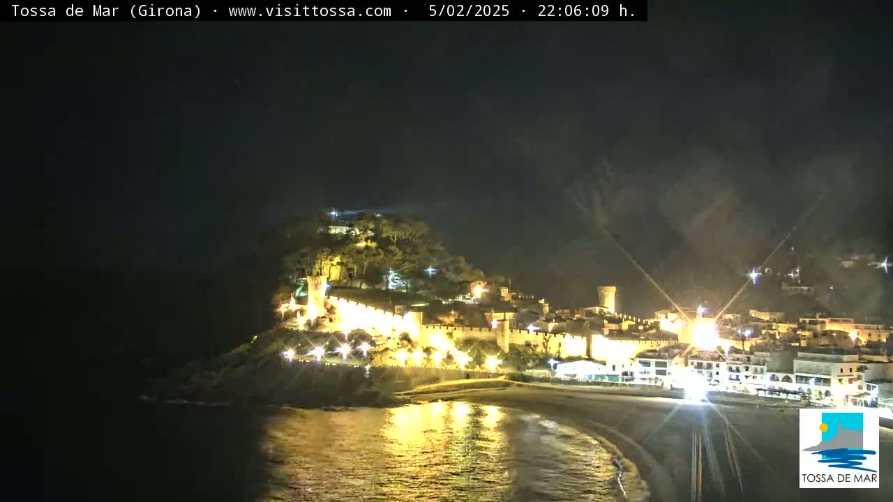 A nighttime view of a coastal town with a medieval-looking castle illuminated on a hill overlooking a dark sandy beach and calm water.