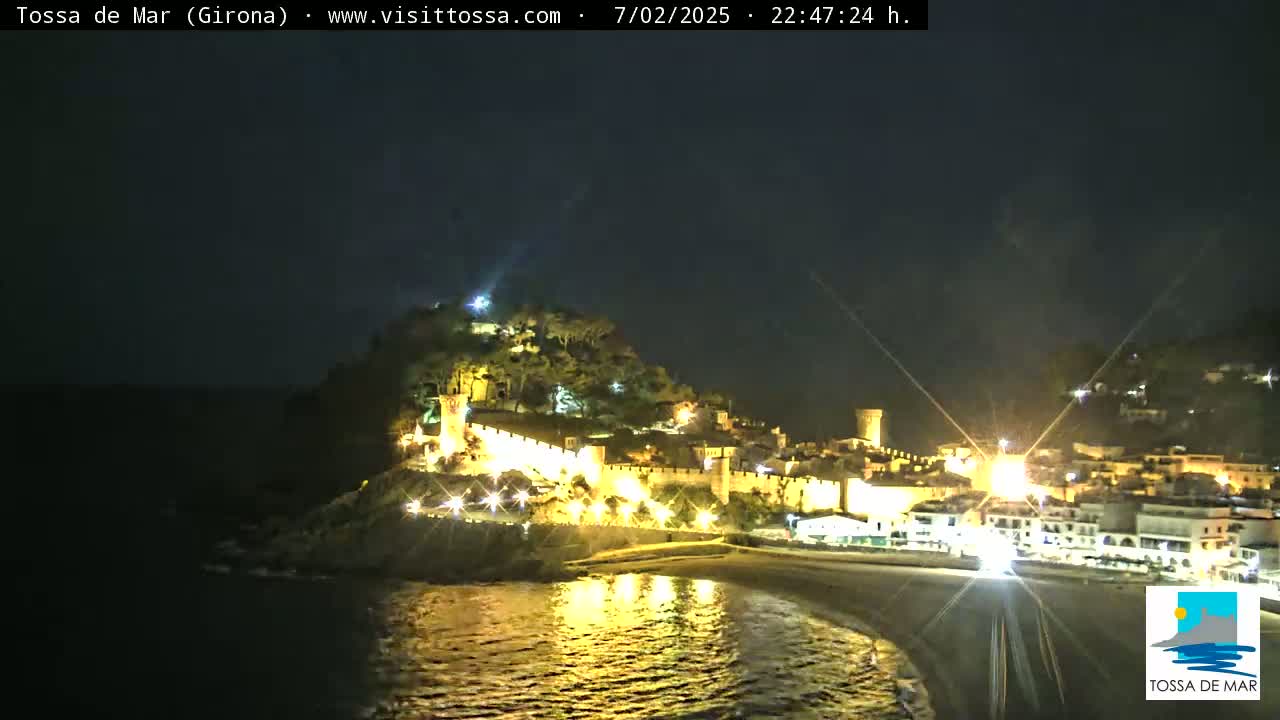 A nighttime view of a coastal town with a walled fortress illuminated on a hill overlooking a beach and calm sea under a clear, dark sky.