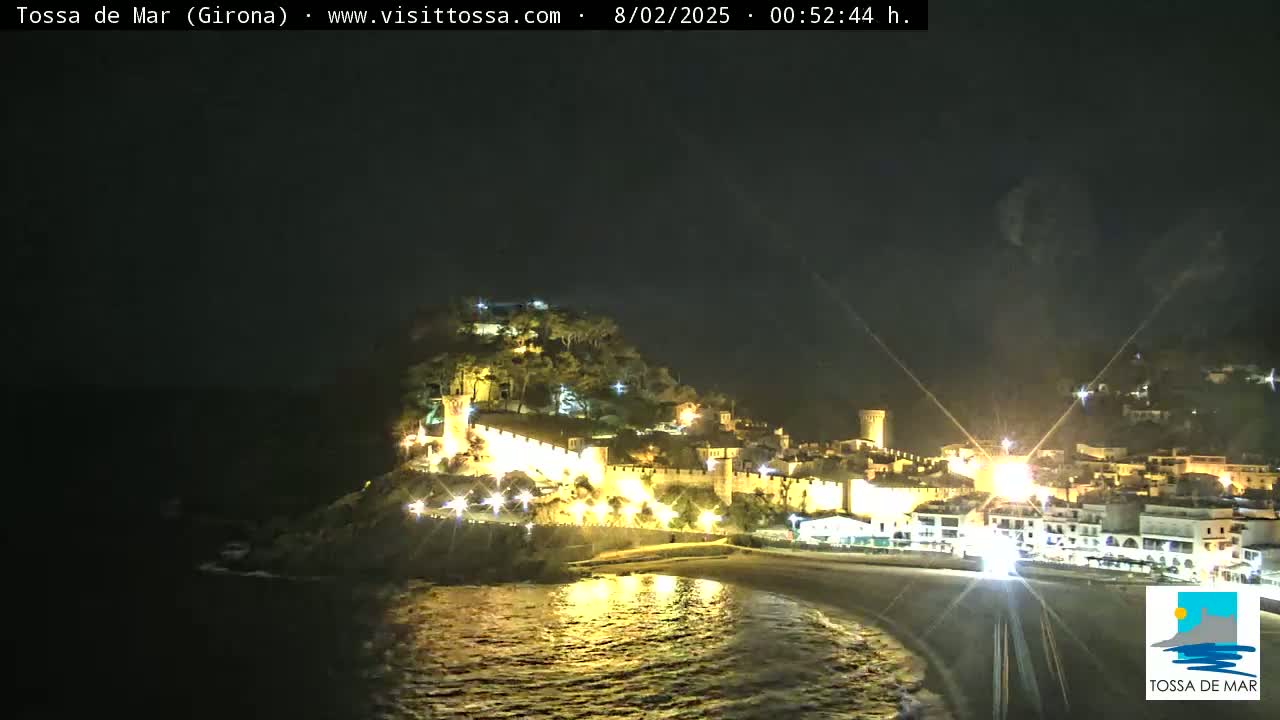 A nighttime coastal town with illuminated buildings and walls sits on a hill overlooking a dark beach and calm water.