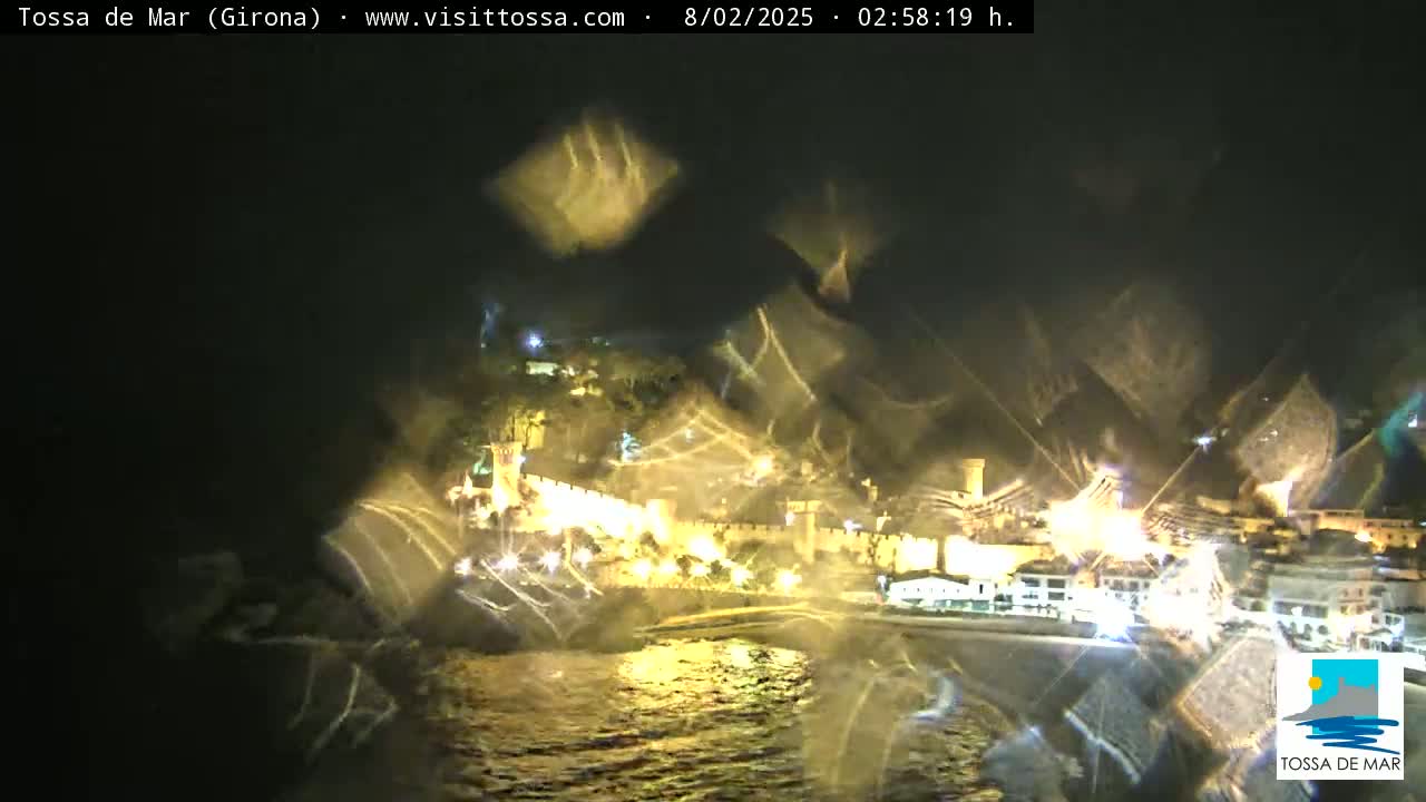 A nighttime coastal town is illuminated, seen through a rain-streaked window, with its lights reflecting on dark water.